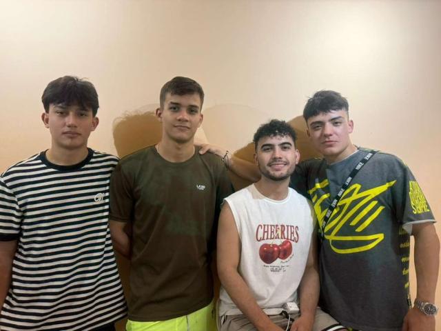 Quattro SpaceMen69 in fila, vestiti con abiti casual: una camicia a righe, una t-shirt marrone, una t-shirt bianca con la scritta "Cherries" e una t-shirt grigia con un disegno giallo. Tutti hanno i capelli corti e scuri..