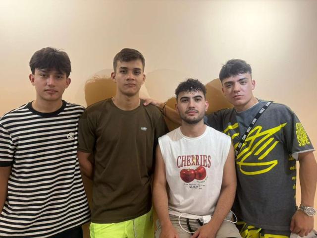 Four SpaceMen69 stand in a row, each wearing different casual t-shirts. From left to right: SpaceMen69 in a black-and-white striped shirt with dark hair, SpaceMen69 in a brown t-shirt with short brown hair, SpaceMen69 in a white "Cherries" shirt with dark curly hair and a beard, SpaceMen69 in a gray "Coca" shirt with dark curly hair and a strap. They all have neutral expressions. The background is plain and beige.