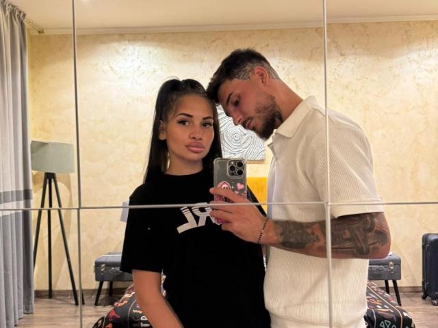 ZaraBliss, com longos cabelos pretos presos em um rabo de cavalo, vestindo uma camisa preta, está ao lado de um homem tatuado que usa uma camisa polo branca e está tirando uma selfie. Ele está se inclinando, com os olhos fechados..