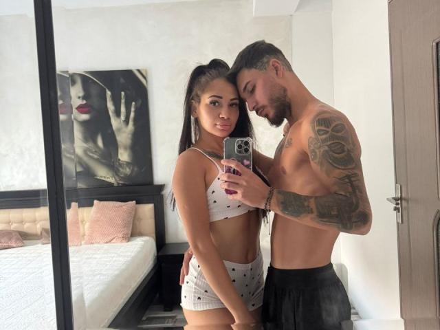 ZaraBliss mit langem, schwarzem Haar zu einem hohen Pferdeschwanz, trägt einen weißen BH und Shorts mit Punkten und steht neben einem tätowierten Mann ohne Hemd, der schwarze Hosen trägt, und macht ein Selfie..