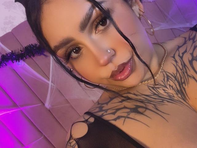 EmmaMyles with black hair, nose piercing, and tattoos, wearing dark lipstick and gold chain, showing off her tits with a mesh top, and a cute smile.