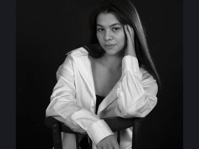 AlmaWhite69 with long dark hair, wearing an open white shirt, resting her head on her hand, sitting on a chair, black and white photo, looking at the camera.