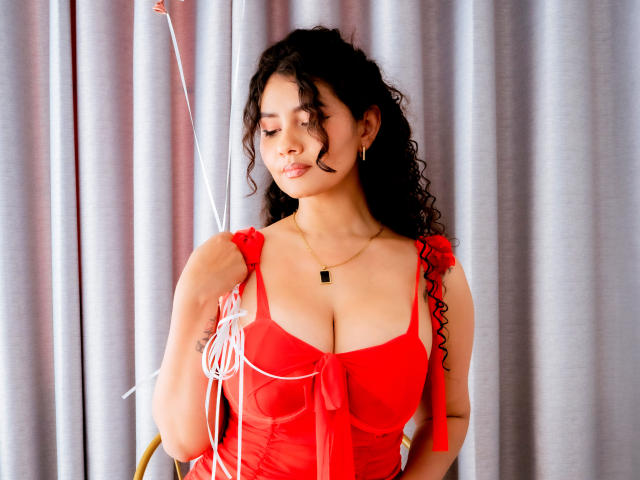 CanelaLebrad with curly black hair, wearing a red dress, holding white ribbons, showing her big boobs, and a black necklace, standing in front of gray curtains.