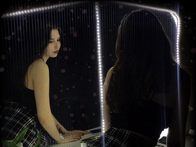 AliceLovels with pale skin, black top, plaid skirt, and red lipstick, leaning on a reflective surface with LED strip lights, long dark hair, tattoo on right forearm.