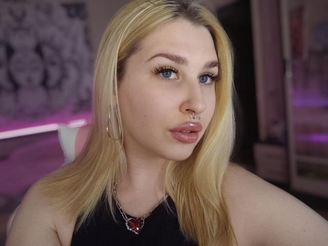 AliceLovels with blonde hair, blue eyes, and a nose ring, wearing a black top and a red heart necklace, looking at the camera with pouty pink lips.
