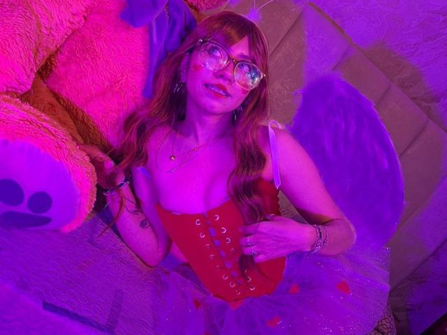 VioletXBloom with long, wavy red hair and clear glasses, wearing a red lace-up corset, lying on a beige couch surrounded by a large pink stuffed teddy bear and purple wings. Purple and pink lighting casts a vibrant glow. VioletXBloom's left hand rests on her corset, right hand on the couch. Her face has a slight smile, visible tattoos on her right arm. The image has a playful, fantasy-like vibe.