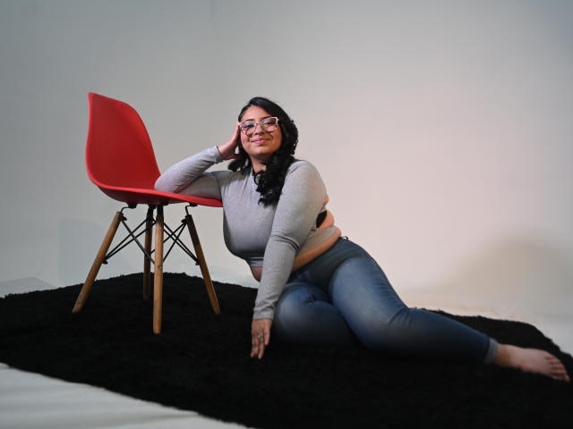 CherylSweetX with glasses, black curly hair, gray crop top, blue jeans, lying on black rug, hand on red chair, smiling, soft lighting, wood legs on chair.