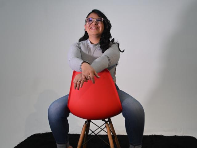 CherylSweetX with glasses, gray long-sleeve shirt, blue jeans, sitting on a red Eames chair, legs spread, smiling, hands on chair, black rug, wood legs, white wall.