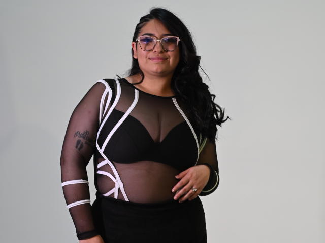 CherylSweetX, a curvy woman with black hair, glasses, and a tattoo on her arm, wearing a sheer black top with white stripes, exposing her big tits.