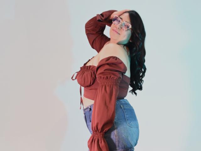 CherylSweetX with glasses, dark curly hair, off-shoulder brown top, showing boobs, blue high-waisted jeans, hand in hair, smiling, light blue background, standing sideways.