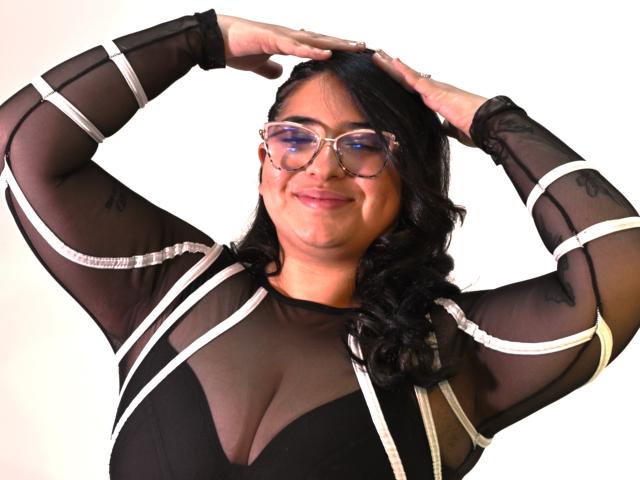 CherylSweetX with glasses, black curly hair, sheer black top with white straps, arms raised, smiling, showing her big tits, white background, looking cute.