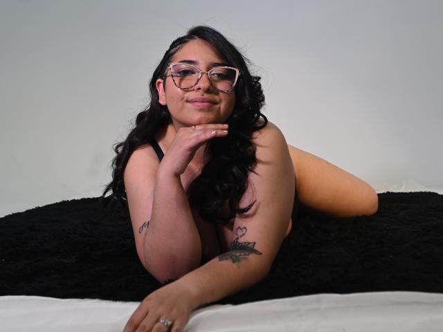 CherylSweetX with long black hair, glasses, and tattoos, laying on a black blanket, smiling, showing her bare tits and ass, wearing a ring on her right hand.