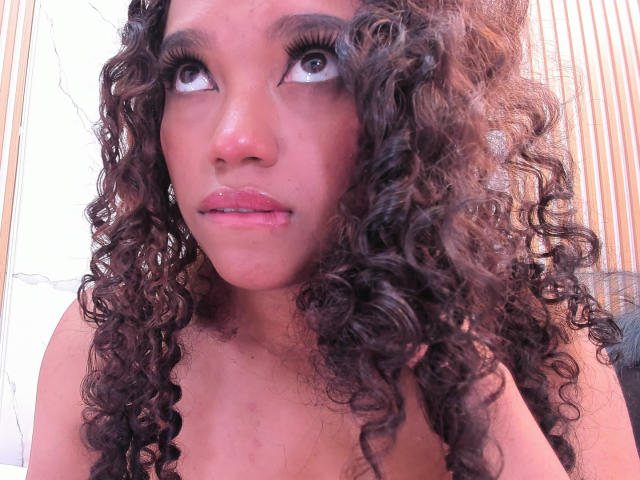 KellyAmbaris has curly brown hair, brown eyes, and pink lips. She's looking up with a slight pout, showing her bare shoulders and a small nose ring.