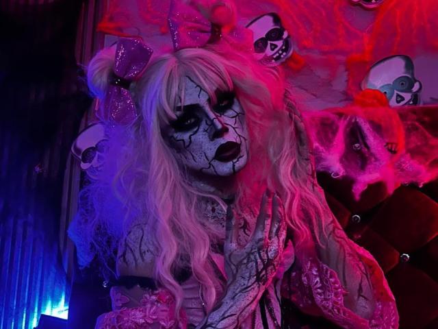 MeganRodriguez in a Day of the Dead costume with white hair, black face paint, pink bow, skull headbands, and cracked makeup. Her hands are raised, fingers spread.