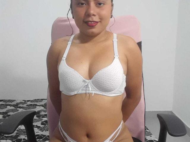 Tretner with medium tan skin, small pink lips, wearing white bra with polka dots and matching white thong, sitting on pink chair, hands behind back, slight smile.