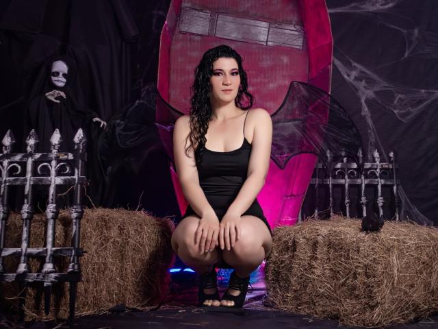 YessiSoto, squatting, wearing a black, tight, spaghetti-strap dress, showing her tits, with wet, curly black hair, in front of hay bales and a skeleton.