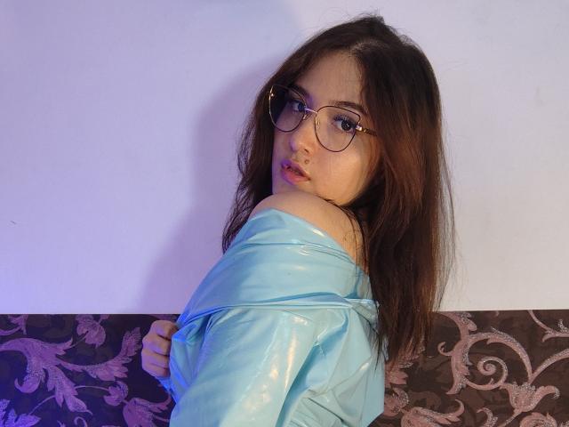 AmaruBloom with long brown hair, glasses, and blue off-shoulder shirt, looking over shoulder, showing a bit of her ass, in a subtle purple-blue light.