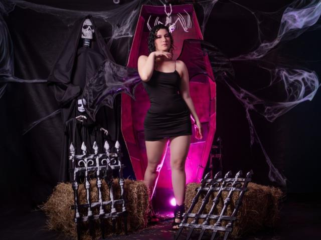 YessiSoto stands in a black dress, holding a pink whip, with a death mask and skeleton in front, hay bales, and a pink coffin behind her.