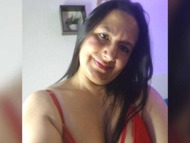 AntonellaGoldy with long black hair, red bra, smiling slightly, medium tits, light brown skin, in a casual selfie. She's leaning forward, looking at the camera.