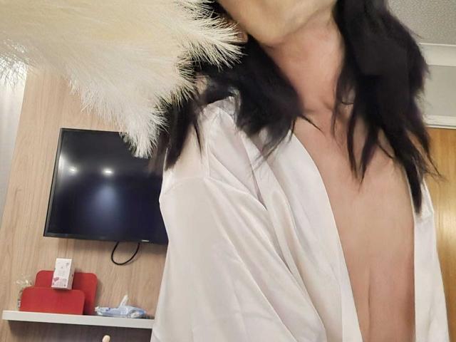 HornyGay69 with black hair, wearing a white, partially open, fluffy-Trimmed jacket, exposing their tits. Flat chest, visible collarbone, and a slightly blurred face. Indoor, casual setting.