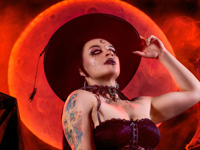 KatalinaJohnson with dark makeup, tattoos, and a witch hat, holding it up, showing off her big tits and chain choker, against a red moon.