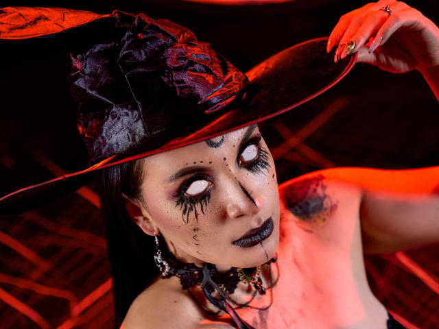 KatalinaJohnson with dark makeup, black lipstick, and freckles, wearing a black witch hat, holding it with red nails, has pale skin and black rose on her head.