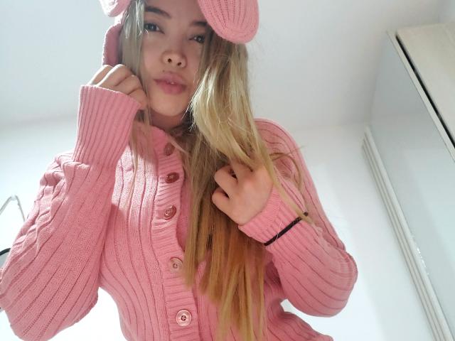 SalomeKiki69 with blonde hair, pink knit hat, and pink sweater, looking down at camera with a subtle smile, showing her pouty lips and cute face.