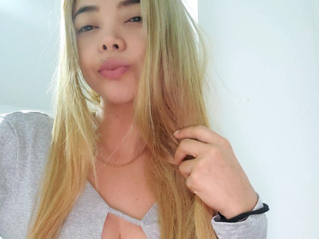 SalomeKiki69 with long blonde hair, gray top, and small tits, holding hair, slight smile, light skin, subtle makeup, wearing a thin gold necklace, black wristband.