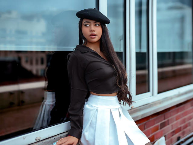 AnnyForger met lang donker haar, een zwarte baret, een zwart topje, een witte plooirok, staat bij een glazen raam, waarbij haar en een bakstenen muur worden weergegeven. Haar borsten zijn klein, haar achterwerk is schattig..