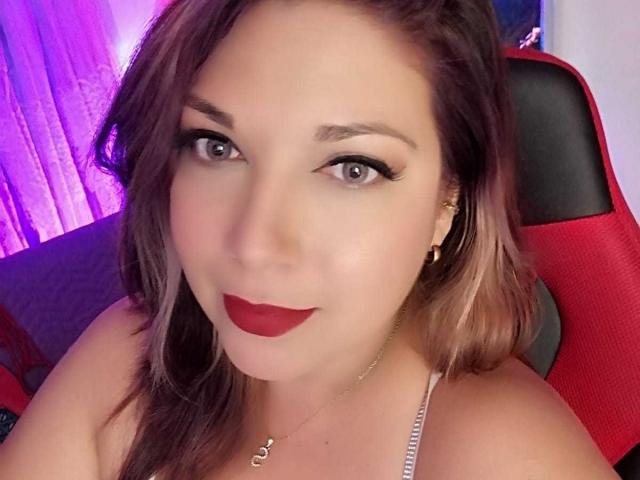 ScarletteThoms has red lipstick, brown eyes with black eyeliner, and light brown hair, sitting in a red chair, wearing a white top, with gold earrings and a necklace.