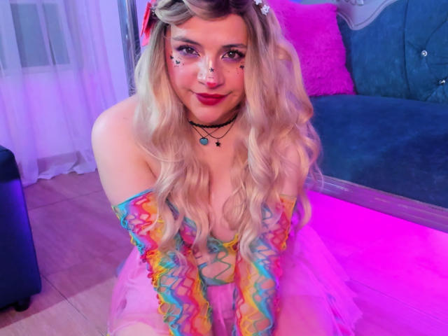 TaraTaylor with blonde wavy hair, colorful off-shoulder dress, pink makeup, and necklace, sitting on pink-lit floor, showing her tits, cute, smiling, in cozy room.