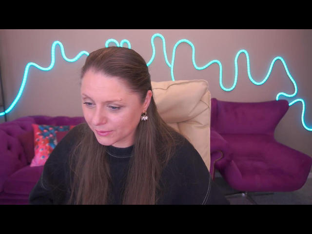 HillsLady has long brown hair, wearing a black top, sitting in a chair with her head slightly tilted down, looking to the side, with a neon blue wire light behind.