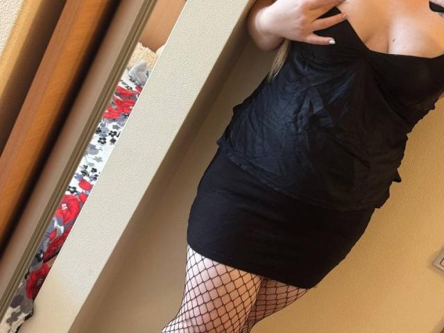 Leiliia in a black, strapless, ruched dress, showing her tits and wearing fishnet stockings, standing in front of a mirror, with her hand near her chest.