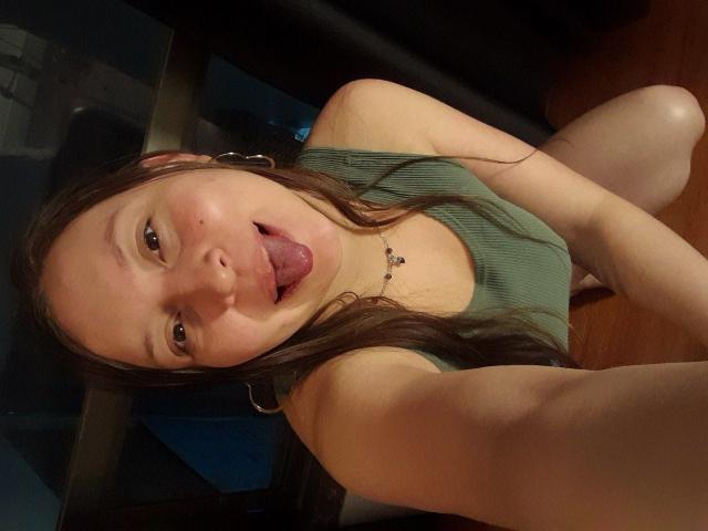 WidnyBeaumont is a cute girl with long brown hair, green tank top, and hoop earrings, sticking her tongue out in a selfie. She's on her knees, showing her ass.