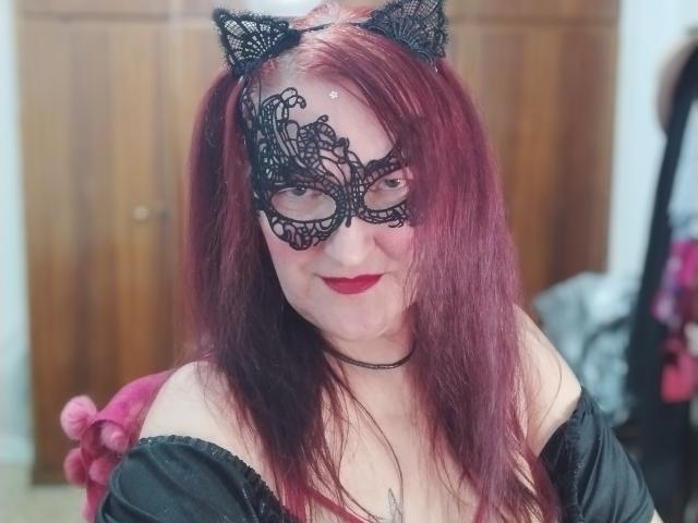 Sabrhina with red hair, black lace mask, and cat ears, smiling with red lipstick, black off-shoulder top, choker, and pink fluffy accessory, showing her tits slightly.