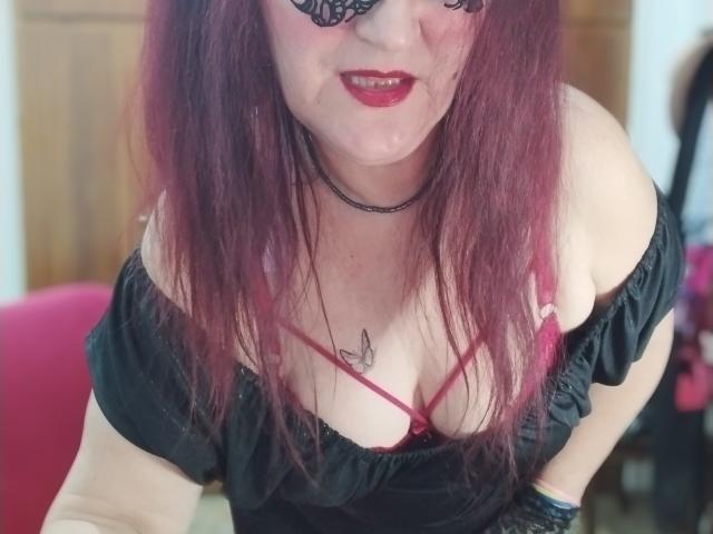 Sabrhina with red hair, black lace mask, red lipstick, black off-shoulder top, showing her tits in a red bra, choker, and tattoo of a bird on her chest.