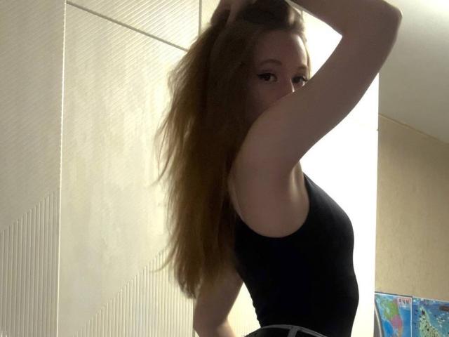 ElfinVibes with long brown hair, wearing a black sleeveless top, holding her hair up, looking over her shoulder, showing her bare back and small ass.