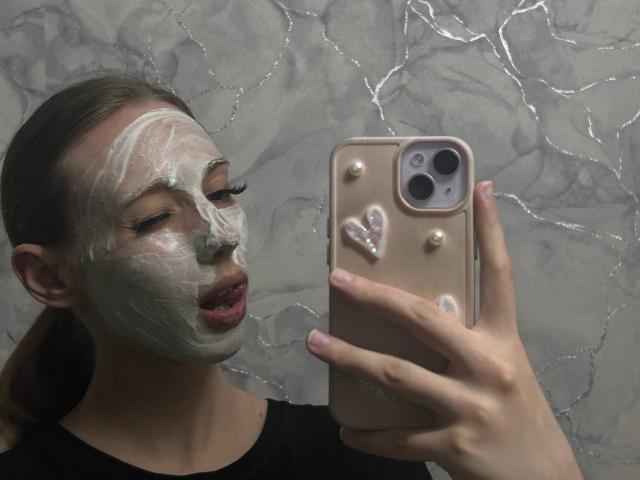 ElfinVibes with a face full of white mask, holding a phone with a pink case and pearl stickers, taking a selfie. Her eyes are closed, and her lips are slightly parted.