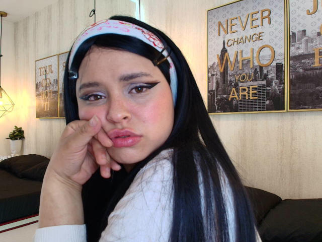 Blueeskyy with long black hair, pink headband, and white makeup, pouting with pink lips, hand on cheek, brown eyes, wearing white shirt, in bedroom with city wall art.