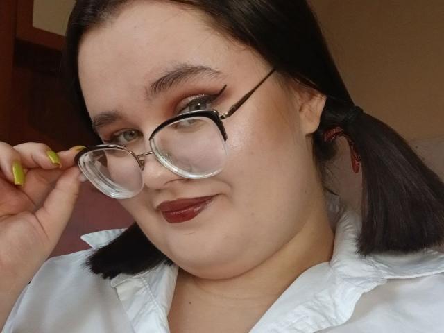 Jessica Joye, con una camicia bianca e voluminosa, due codini neri, unghie verdi, occhiali e rossetto rosso, tiene un bicchiere con la mano destra, ha un sorriso carino e un viso paffuto..