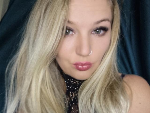 AlmaDeAngel69 has blonde hair, green eyes, a nose ring, and pink lipstick. She's wearing a black, studded top. Her pouty lips are slightly glossy.