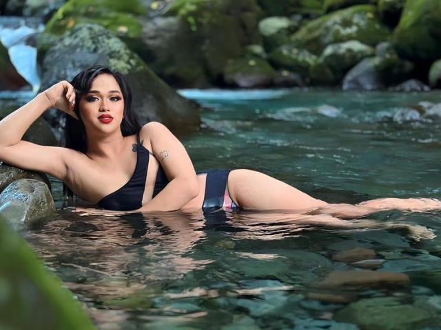 AmaraHugeCock is lounging in a rocky stream, partially submerged in clear water. She has long black hair, red lipstick, and is wearing a black one-shoulder bikini with a pink waistband. Her right arm rests on a rock, and her left arm is bent, with her hand near her head. Her skin is light, and she has a small tattoo on her right arm. The background includes moss-covered rocks and a small waterfall. The water reflects her image and the rocks. The image has a natural, relaxed vibe.