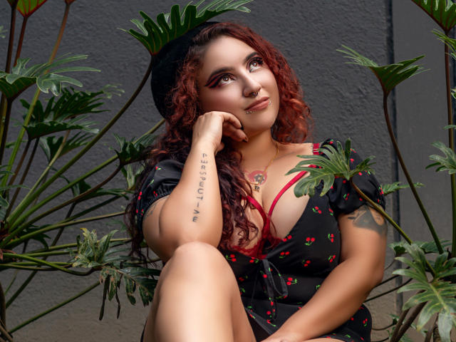 RavenWaifu with curly red hair, black dress with cherry patterns, red bra, tattoo on left arm, black beret, sitting among green leaves, hand on cheek, sexy, confident.