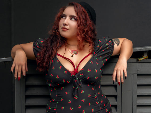 RavenWaifu with red curly hair, black beanie, red bra, cherry-pattern dress, large tits, red nails, black tattoos, leaning on grey metal structure, looking up, confident.