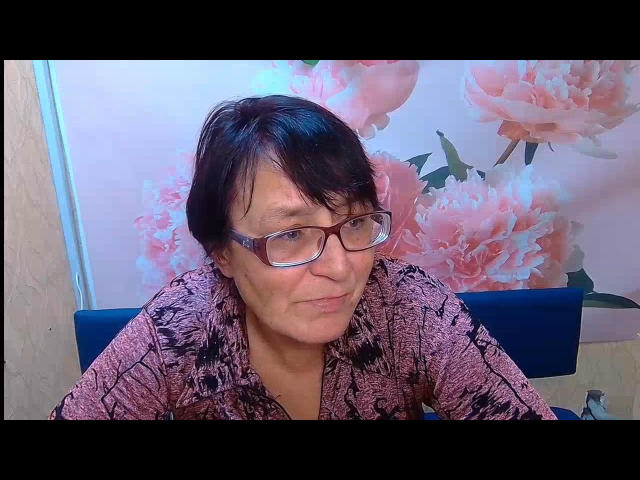 MeganSord with short black hair, glasses, and a pink and black patterned shirt, smiling slightly, sitting in a blue chair. Her tits are small and she's cute.