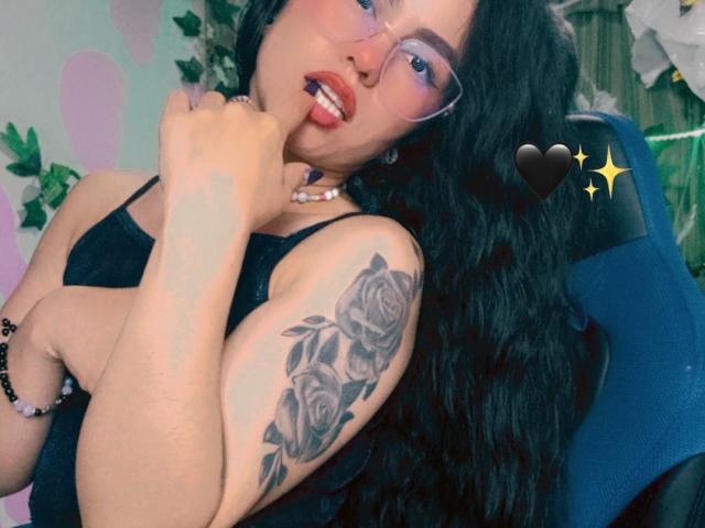 Lucecita69 with long black hair, wearing glasses and a black top, showing a rose tattoo on her arm, biting her finger, and a heart emoji. Her ass is not visible.