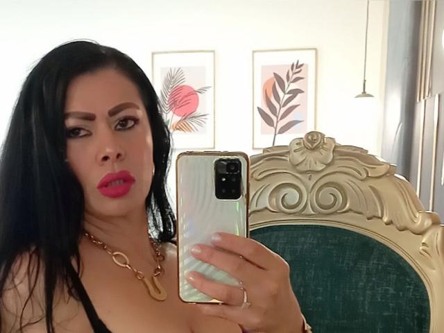 SofiMontes with long black hair, red lipstick, and gold necklace, taking a mirror selfie with her phone, showing her boobs in a green and gold chair.