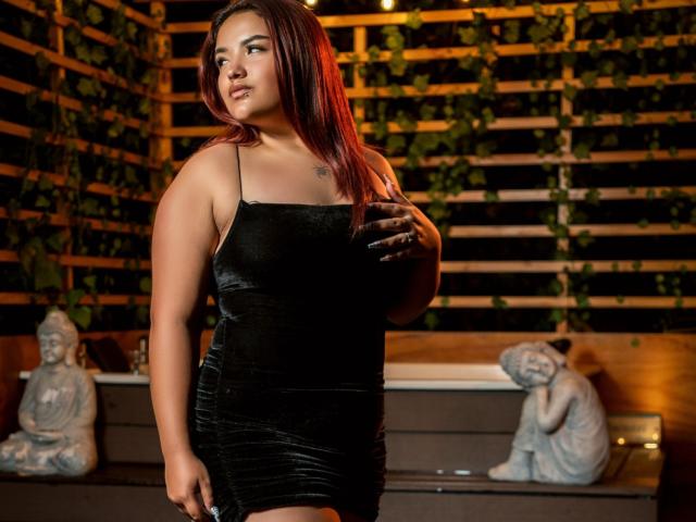 LilithMoonryn, a plus-sized woman with red hair, wears a tight black velvet dress, showing her ass and small tits, standing with a hand on her chest, looking sideways.