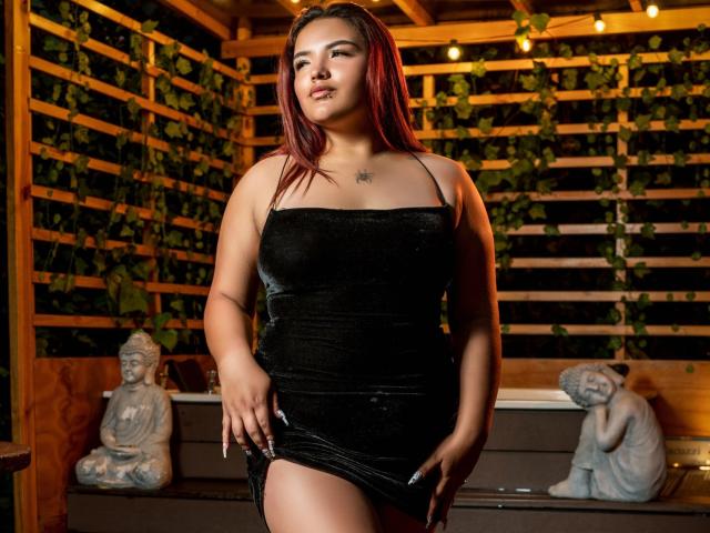 LilithMoonryn with red hair, wearing a tight black velvet dress, showing her ass with a hand on her thigh, standing in a wooden room with statues.