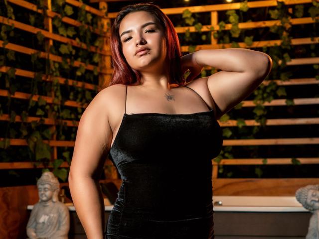 LilithMoonryn with red hair, black velvet dress, and a small tattoo on her chest, hand on her head, looking confident. Her tits are visible, and she's sexy.